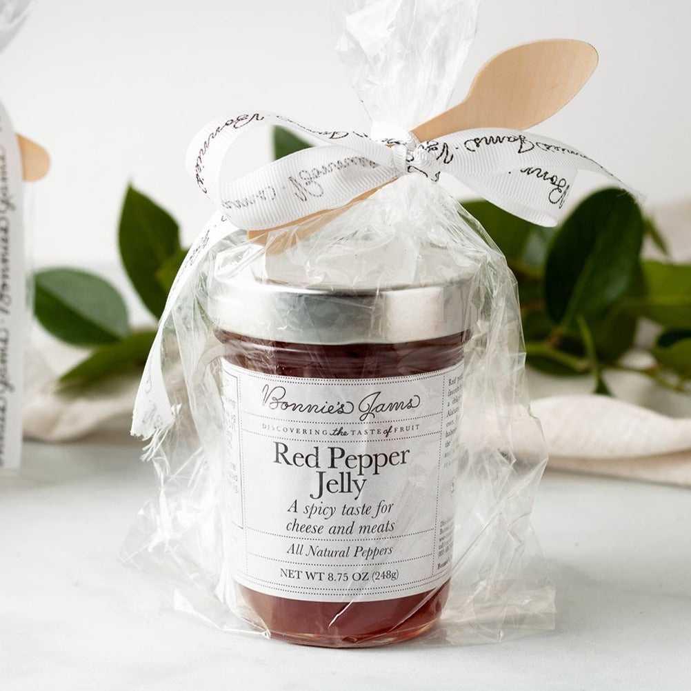 A jar of Red Pepper Jelly in a clear cello bag, with a wooden spoon tucked in the ribbon that ties the top.